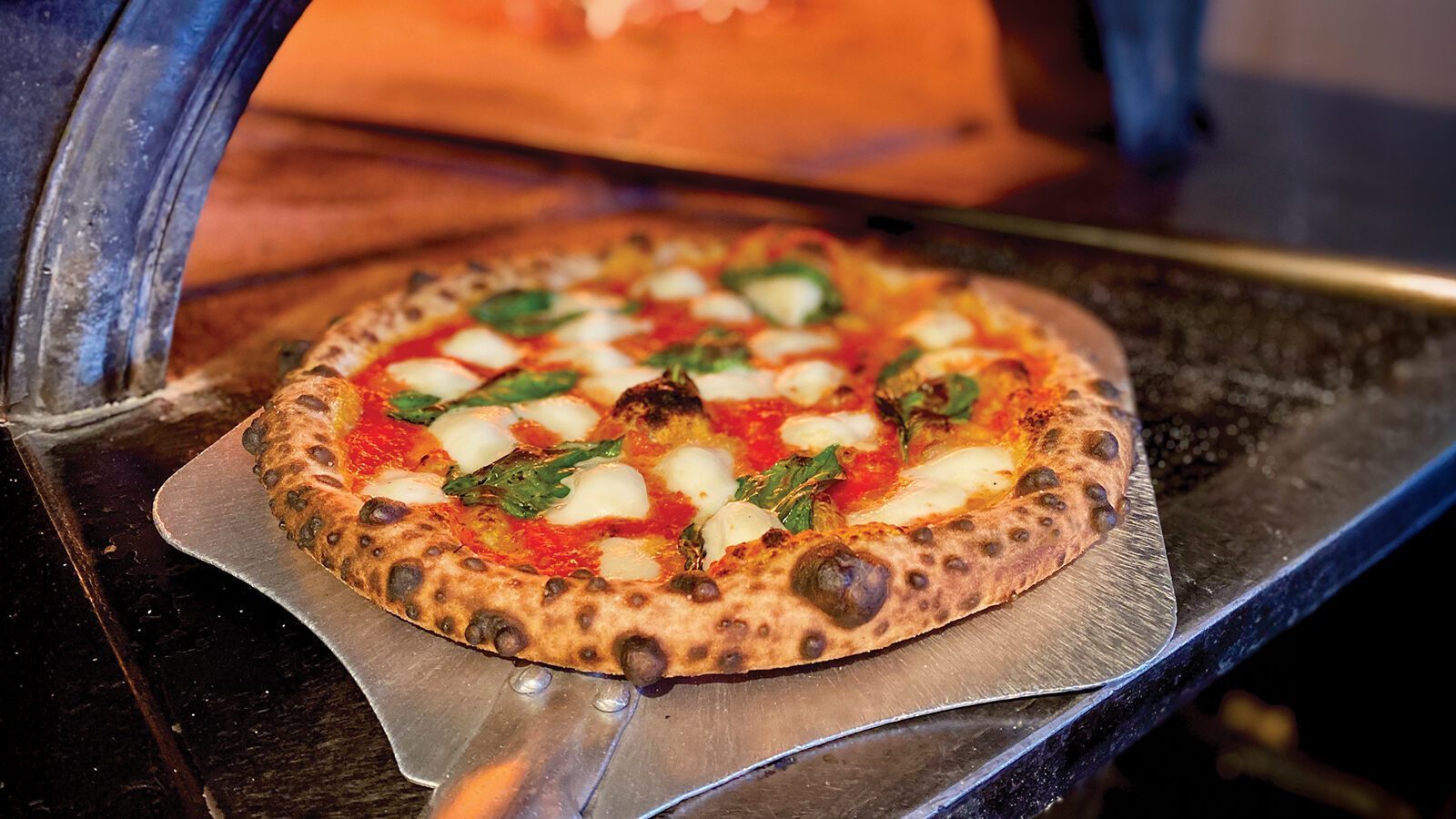 A wood-fired pizza with tomatoes, mozzarella, and basil sits on a metal pizza peel in front of the oven at Bar Del Corso, a favorite spot for pizza lovers in Beacon Hill.