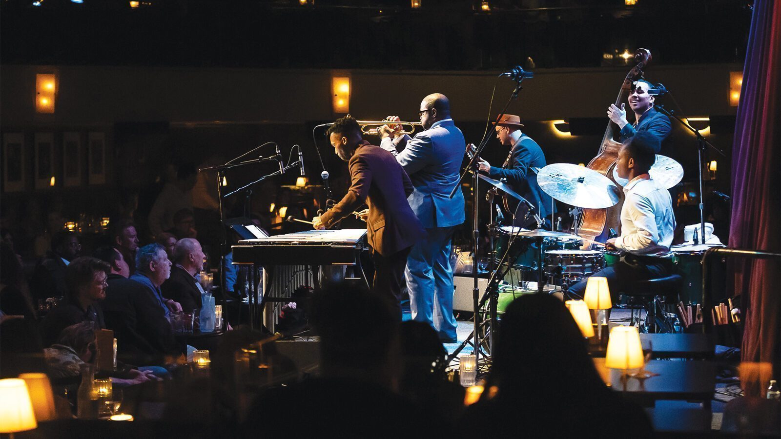 A jazz band performs live music on stage at Dimitriou's Jazz Alley, entertaining a seated audience in a dimly lit jazz club with small tables and glowing lamps.