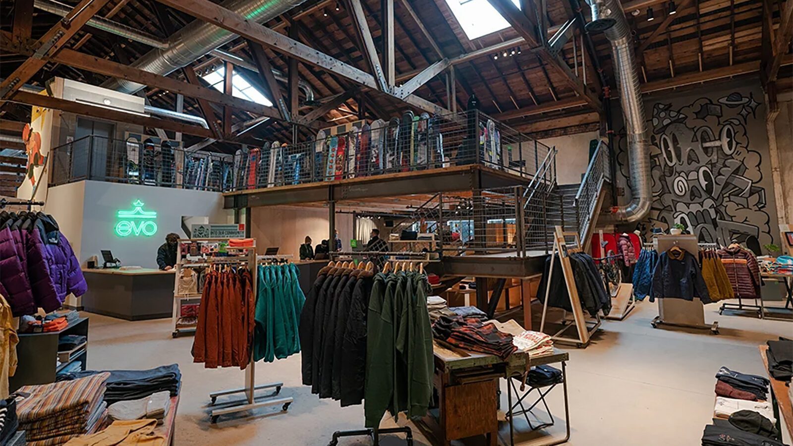 Clothing and outerwear displayed on racks inside a spacious, industrial-style Evo retail store—founded by skier Bryce Phillips—with exposed beams, mezzanine level, and a mural on the wall.