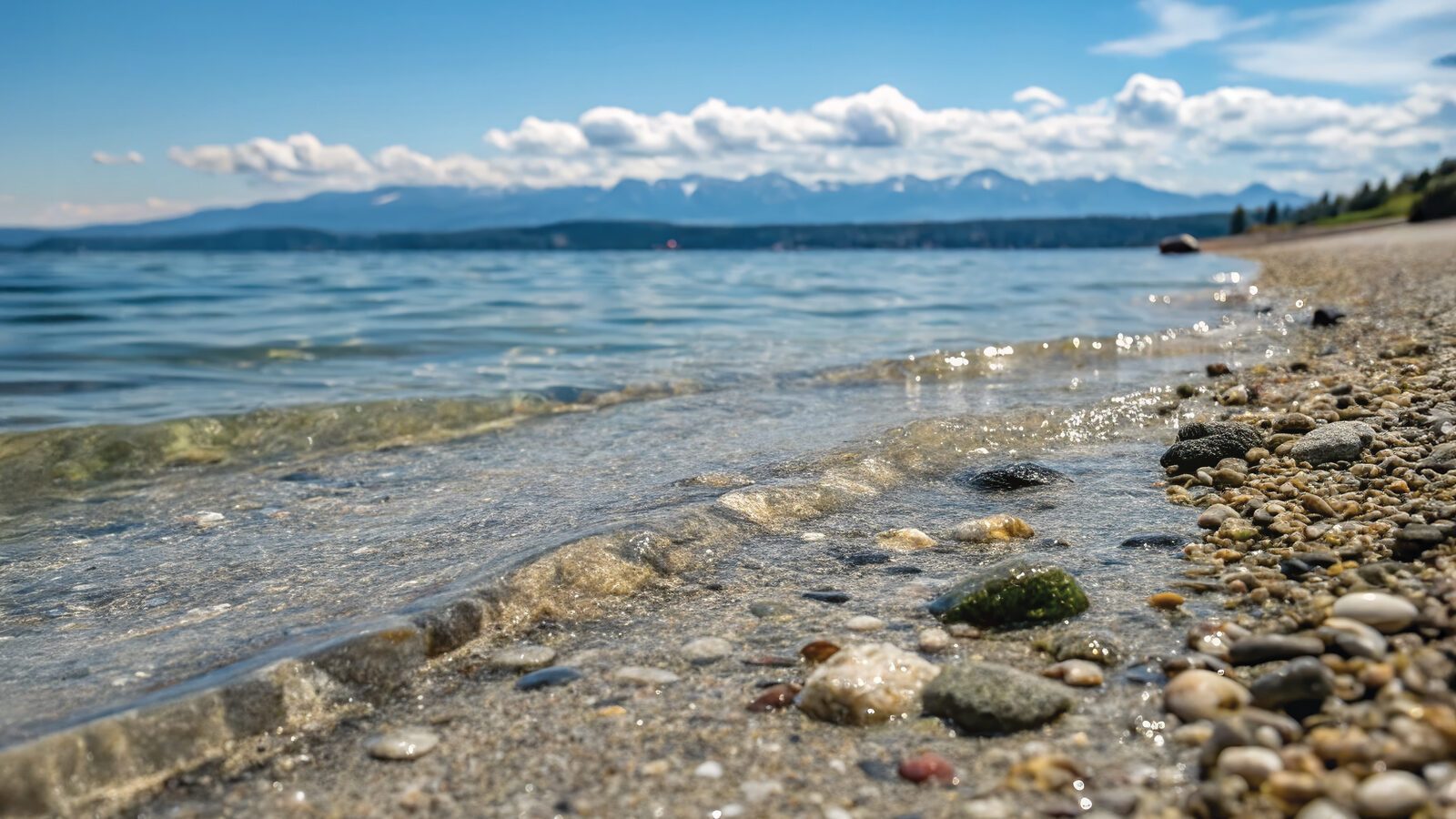Close-up of a pebbled shoreline in a charming beach town, with gentle waves, clear blue water, and distant mountains—a scene worthy of art under a partly cloudy sky.