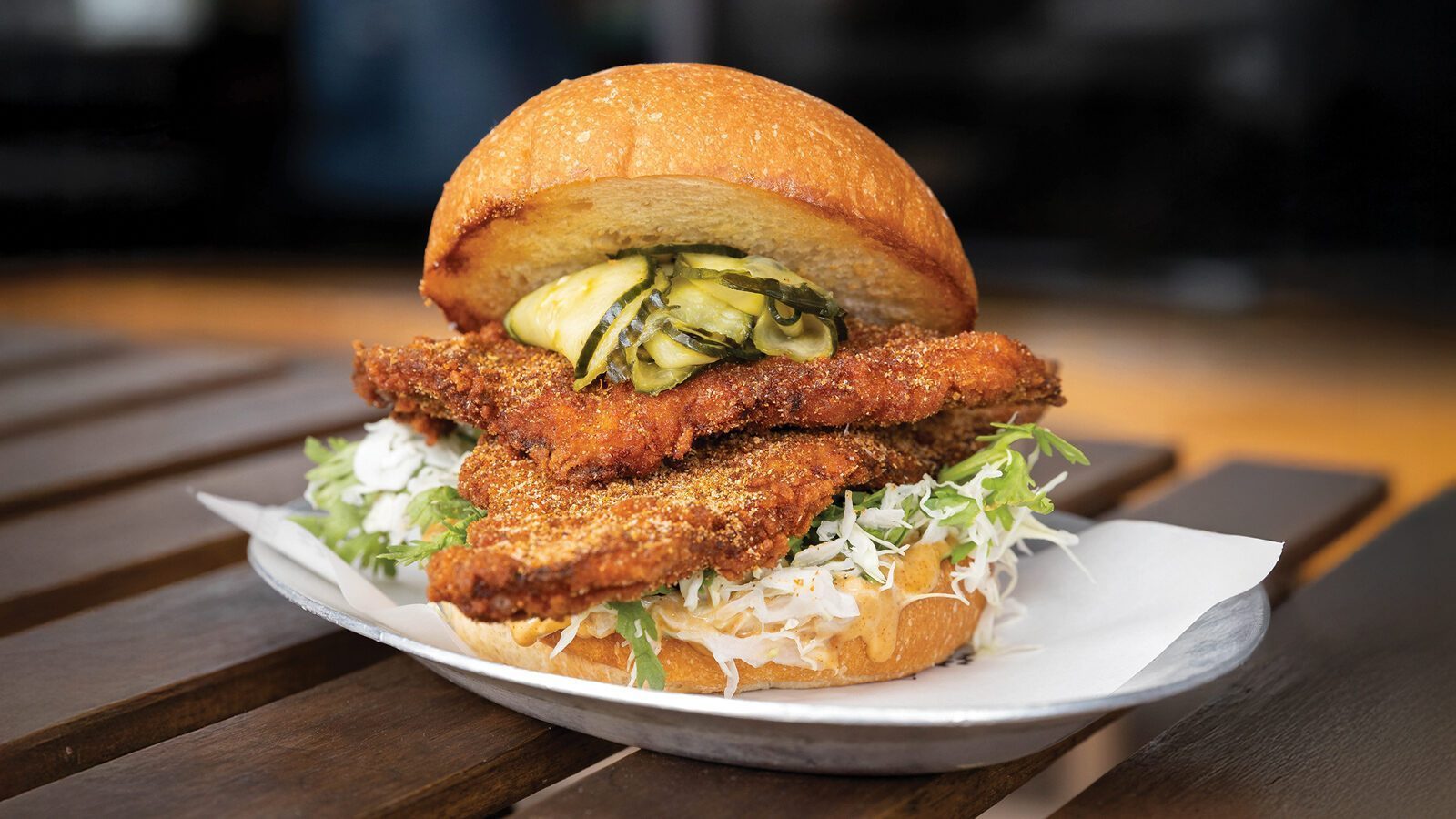 A Local Tide fried chicken sandwich with shredded lettuce, pickles, and a toasted bun is served on a plate lined with paper.