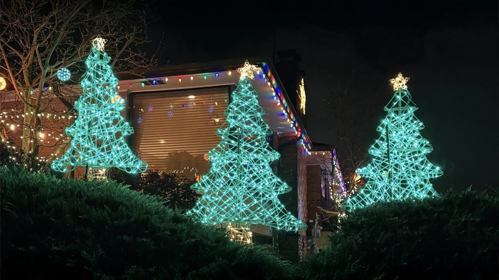 Three glowing wireframe Christmas tree decorations light up a lawn in Olympic Manor, with the house behind them beautifully adorned in multicolored holiday lights for December nights.