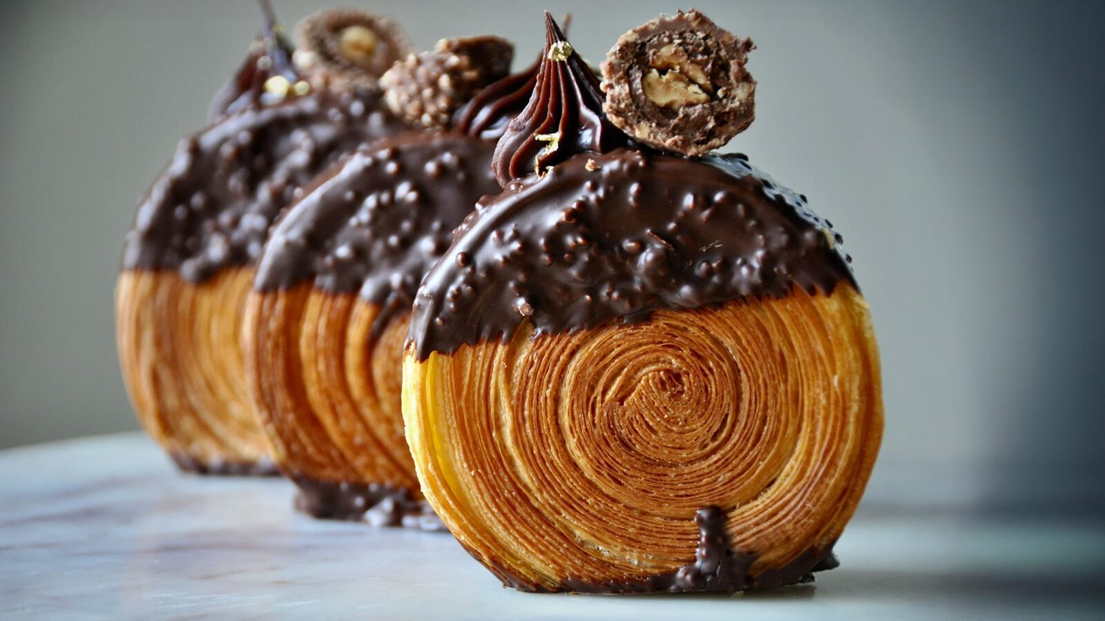 Three chocolate-dipped Petit Pierre pastry rolls with visible flaky layers, topped with a chocolate swirl and a round chocolate candy, arranged in a row on a light surface—truly the Best of the Best.