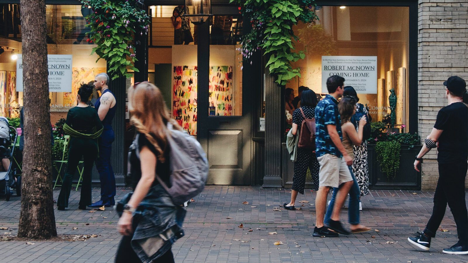 People walk past a Seattle art gallery in Pioneer Square with colorful artworks displayed in the windows; some individuals are talking or looking inside while others move along the sidewalk during the lively art walk.