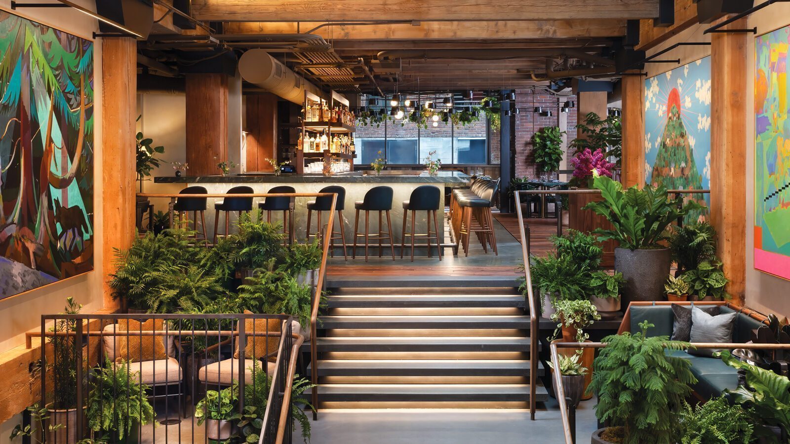 A modern bar and lounge at Populus Seattle with wooden beams, large colorful paintings, abundant green plants, and a central staircase leading to a bar counter with high stools.