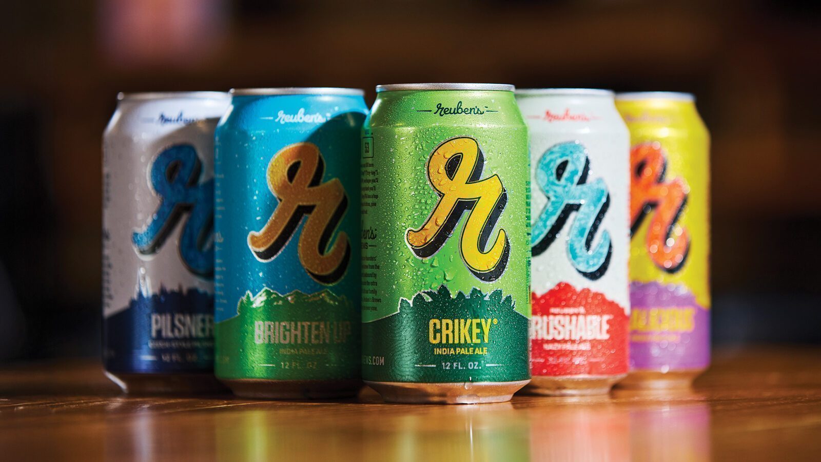 Five cans of Reuben's Brews, a leading craft brewery, are arranged in a row on a wooden surface, each can in a different color and style and covered with refreshing condensation.