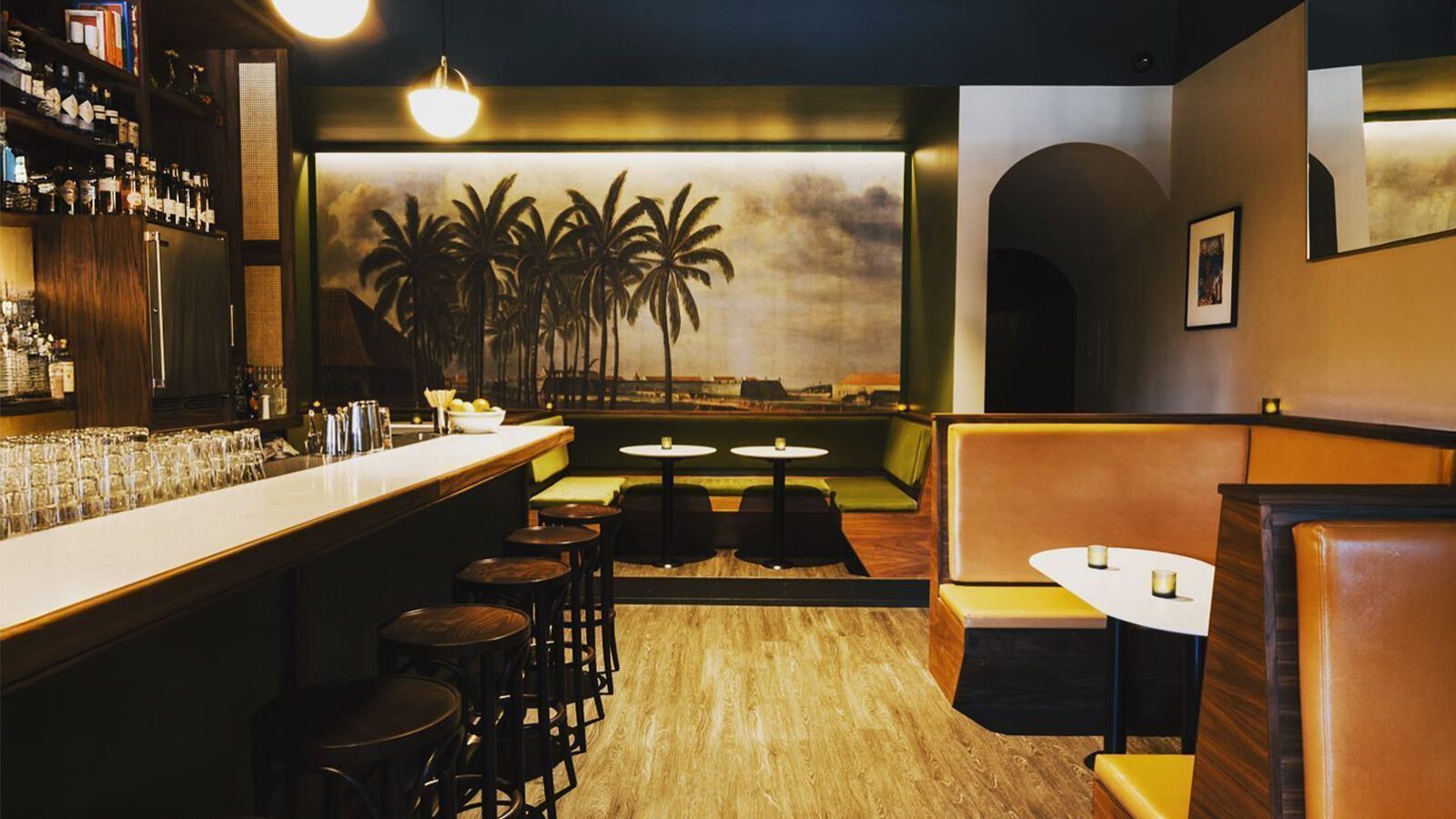 Roquette is a modern craft cocktail bar with a long counter, black stools, brown booths, small tables, and a mural of palm trees on the back wall—perfect for enjoying drinks at this 2025 James Beard Award contender.