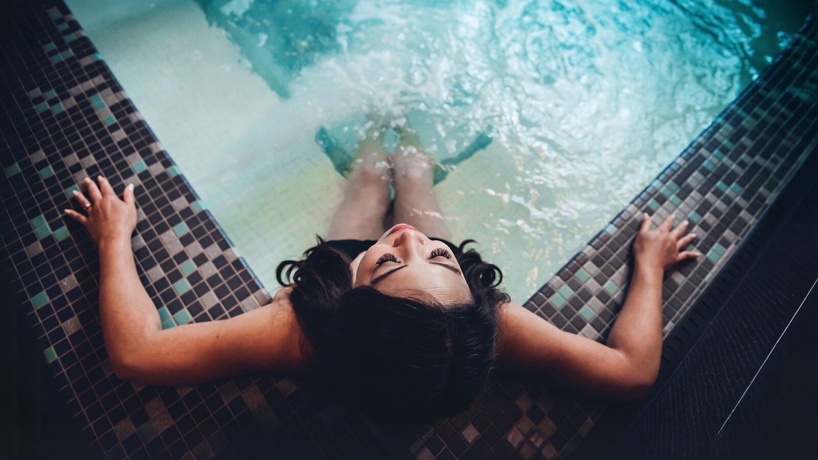 Unwind at Yuan Spa: a person relaxing in a tiled indoor pool, leaning back against the edge with arms outstretched and head tilted upwards, experiencing the best spa tranquility.