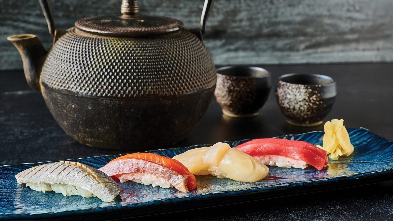 A rectangular plate from Sushi Kashiba with assorted nigiri, pickled ginger, a teapot, and two teacups sits on a dark surface—showcasing some of the best sushi in town.