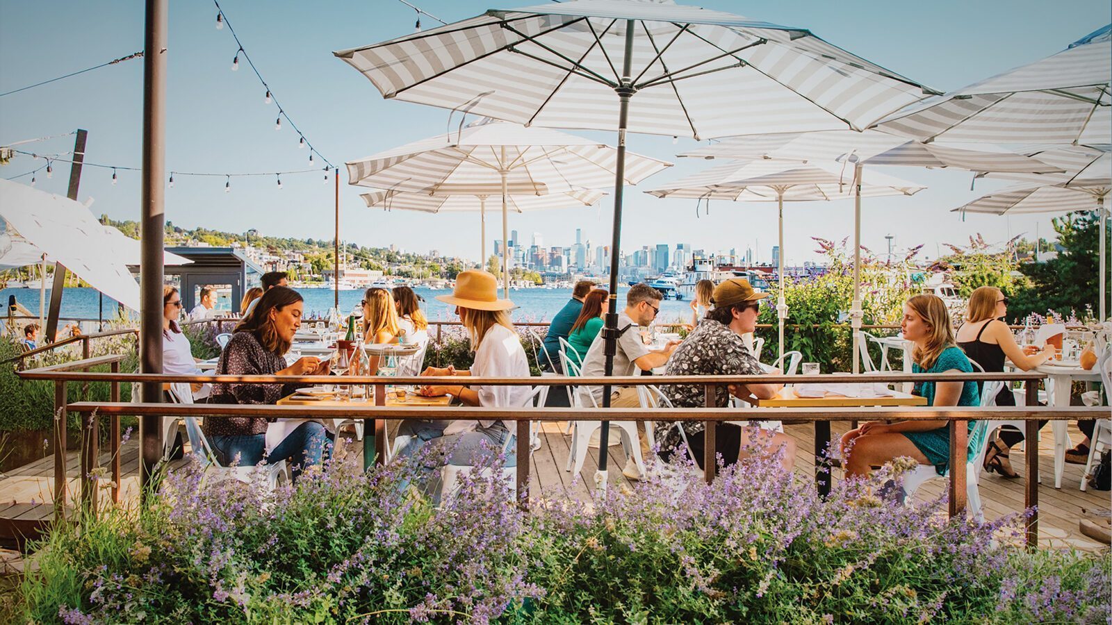 People enjoying outdoor dining at Westward under large umbrellas on a sunny day, with the city skyline and water in the background—a signature experience inspired by chef Renee Erickson.