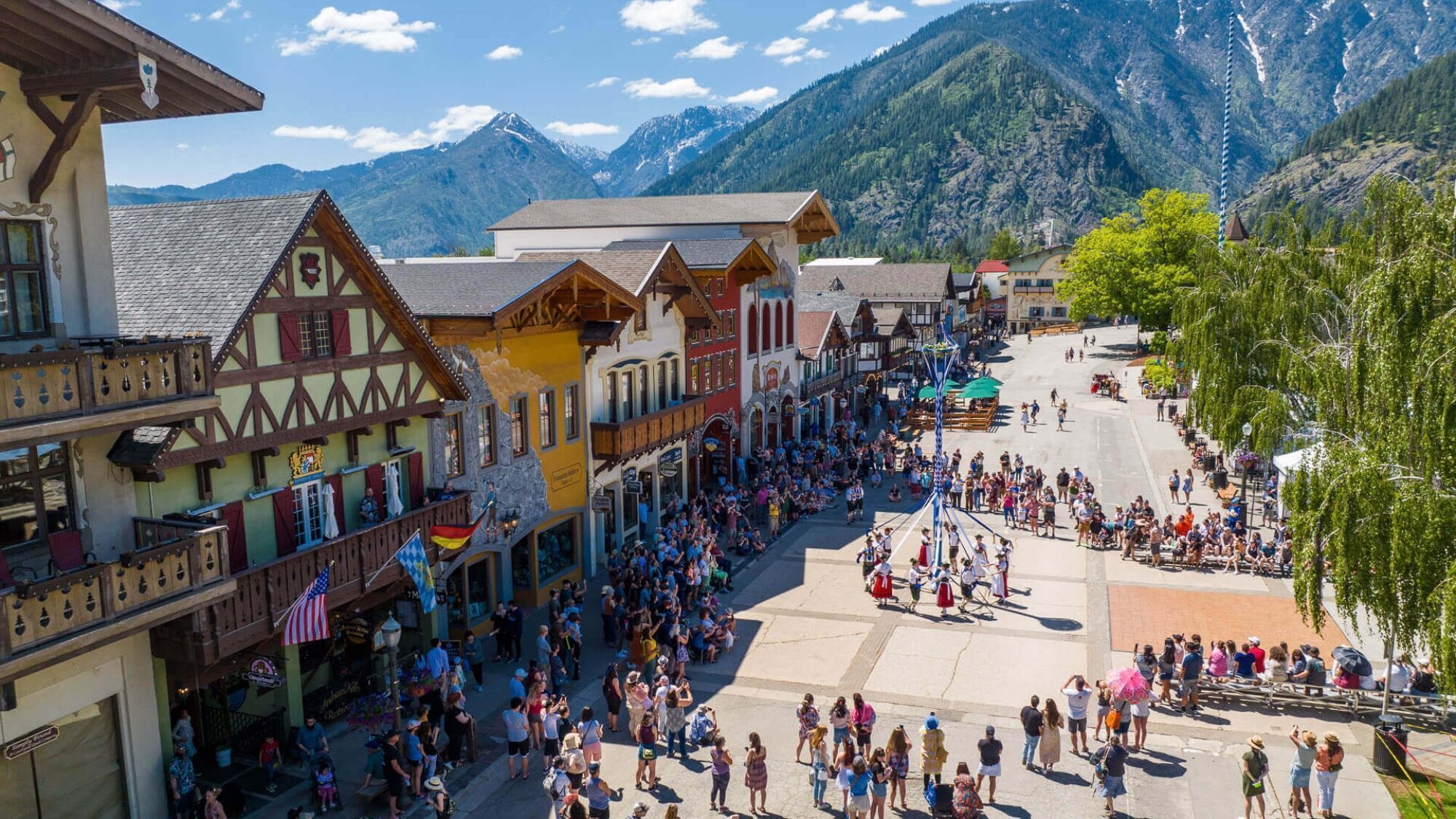 A crowd gathers in a town square with colorful Bavarian-style buildings, set against a backdrop of mountains under a clear blue sky.