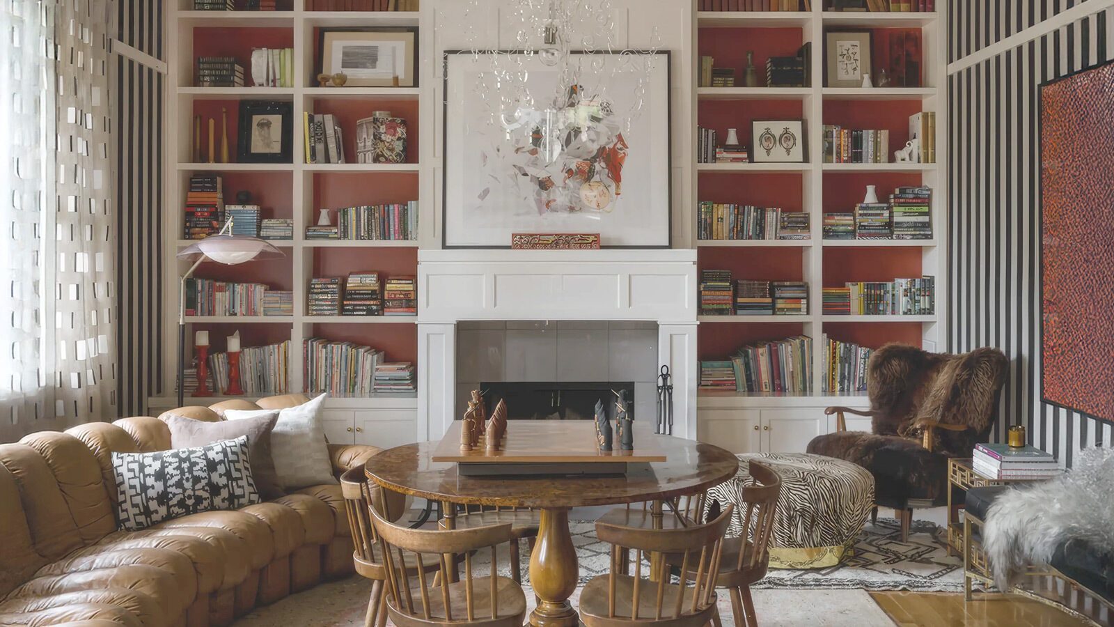 A living room with built-in bookshelves, a fireplace, a round wooden table with chairs, a sectional sofa, and a fur chair under a chandelier.