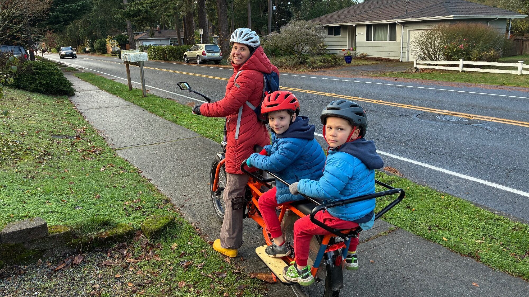 An adult and two children wearing helmets and jackets sit on an electric bike on a residential sidewalk next to a street with houses and trees.