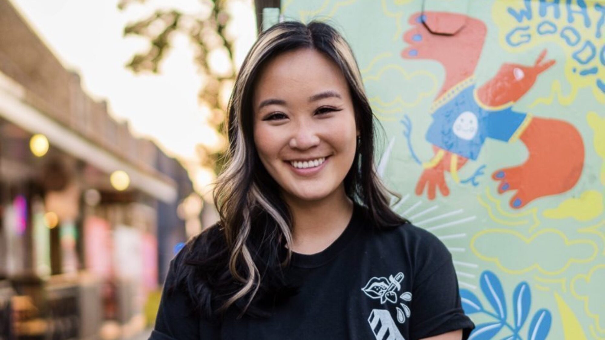 A woman with long hair and a black shirt stands smiling in front of a colorful mural featuring abstract shapes and characters.