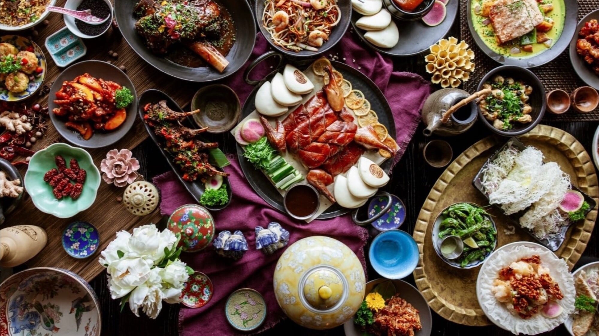 A variety of Chinese dishes, including roast duck, steamed buns, seafood, vegetables, and sauces, are arranged on a table with decorative plates and teapots—perfect for a unique Thanksgiving dining experience.