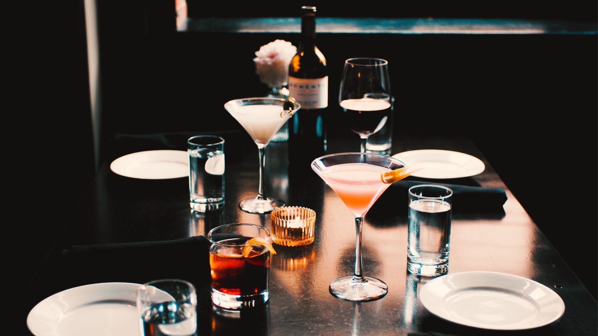 A dark wooden table set with four empty plates, glasses of water, three cocktails, a glass of red wine, a wine bottle, and a candle in the center.