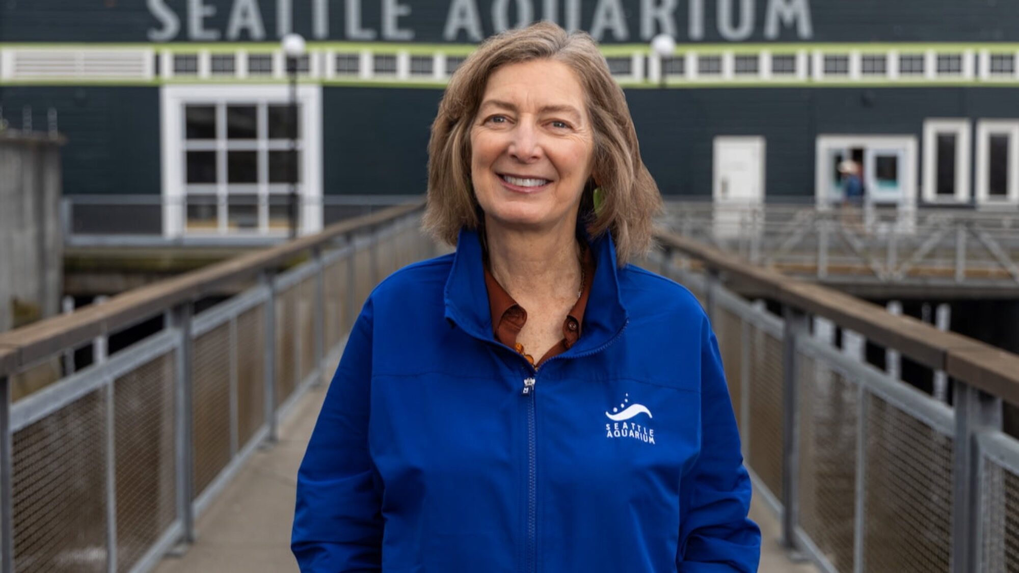 Peggy Sloan, in a blue Seattle Aquarium jacket, stands on a walkway in front of the Seattle Aquarium building, smiling at the camera and celebrating marine-life conservation.