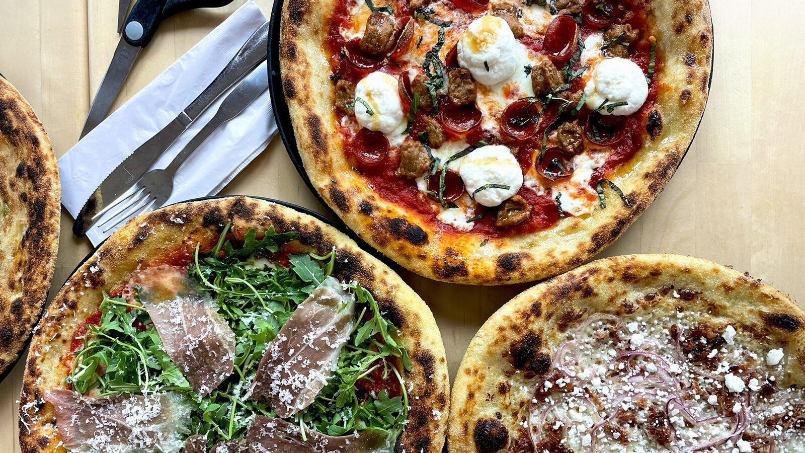 Three wood-fired pizzas from Sourdough Willy’s on a table, topped with arugula and prosciutto, mozzarella and tomatoes, plus cheese with onions. Cutlery and a pizza cutter complete this Counter Culture-inspired feast.