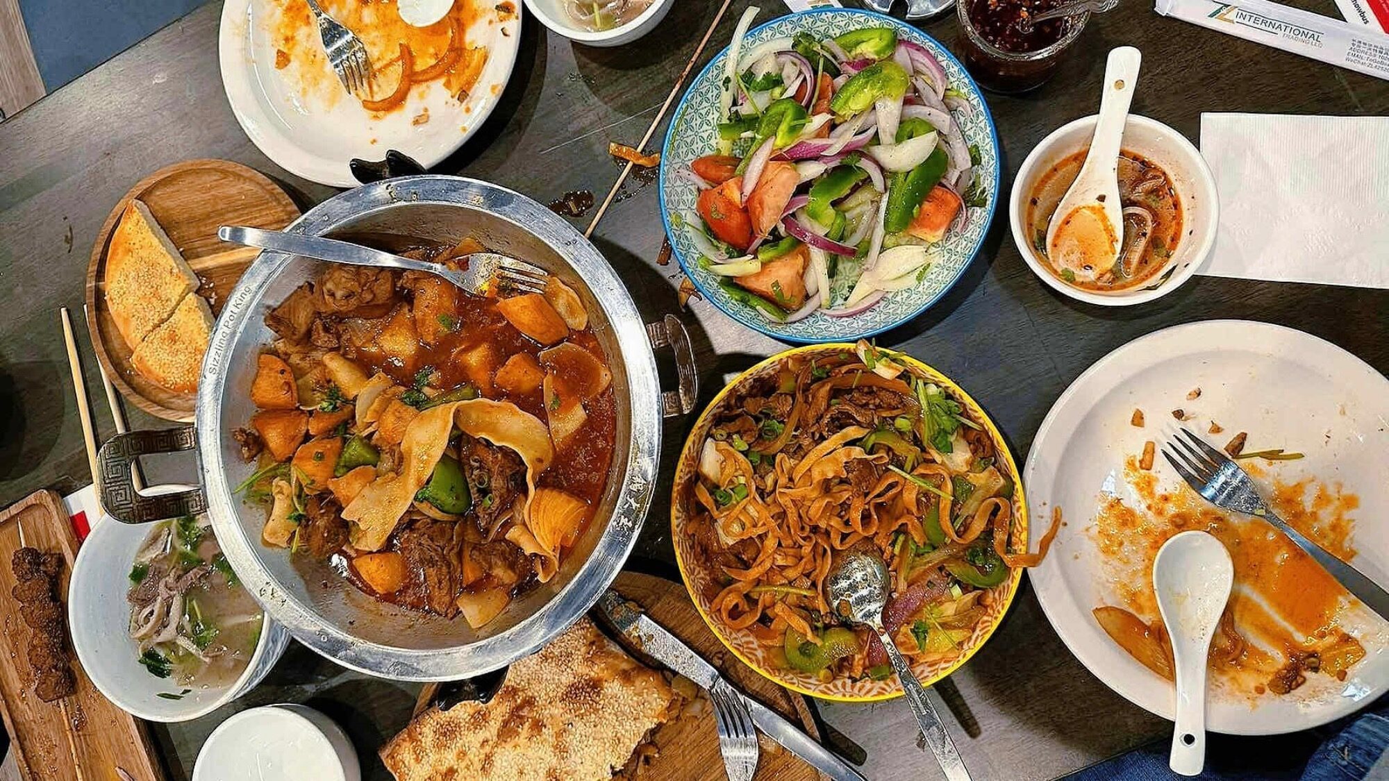 A table with various dishes including a hotpot, noodles, salad, soup, bread, and partially eaten plates with utensils.