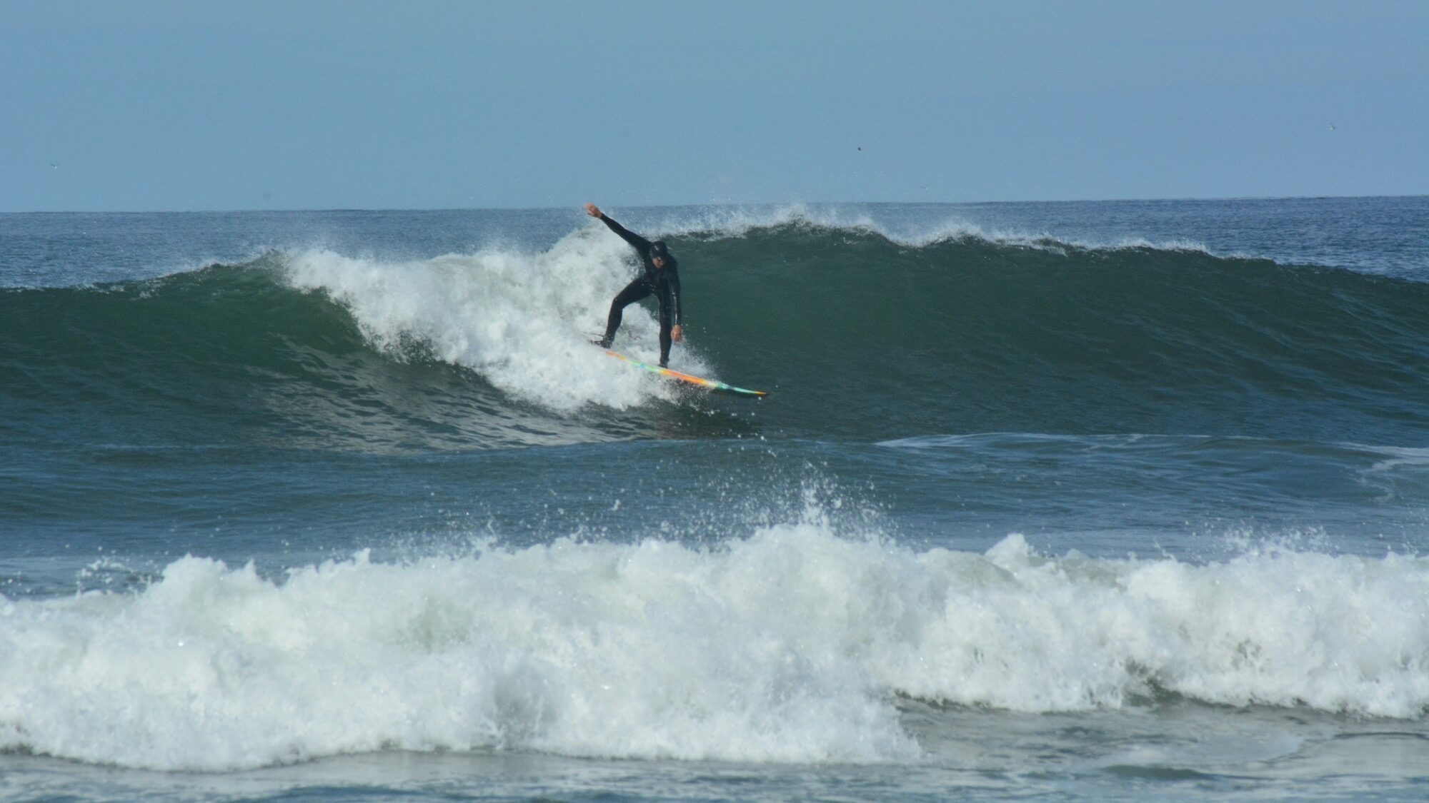 A surfer in a black wetsuit rides a wave on a surfboard in the ocean under a clear sky.