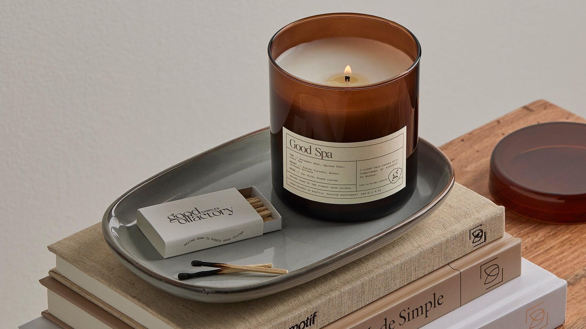 A lit brown glass candle labeled "Good Spa" sits on a gray tray beside a matchbox and matches, atop a stack of books and a wooden surface.