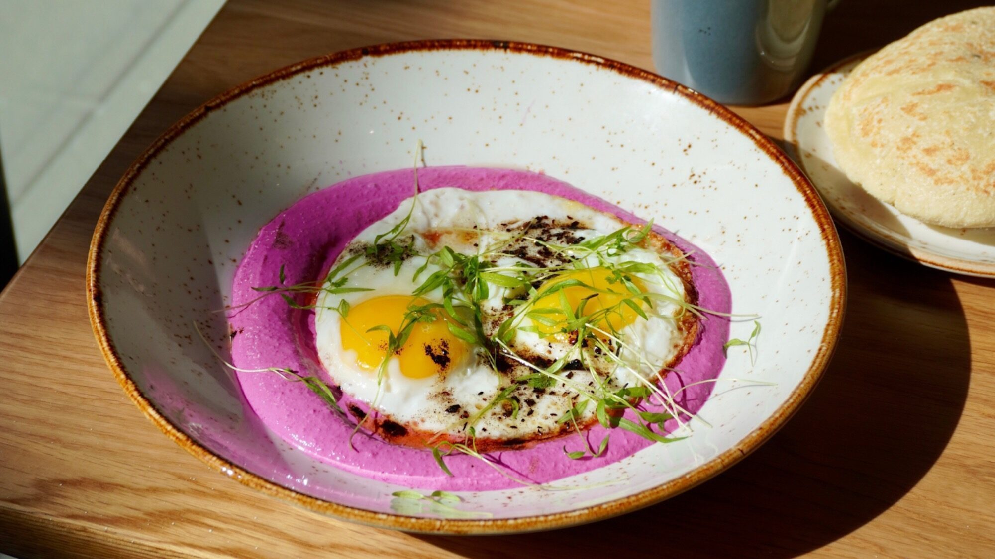 A bowl with two sunny-side-up eggs and microgreens on a layer of purple puree, placed on a wooden table next to a bread roll and a mug.