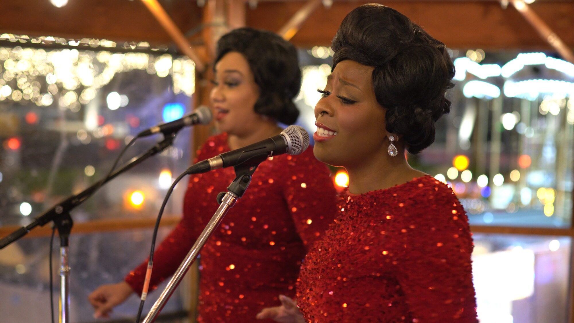 Two women in matching red sequined dresses sing into microphones on stage, with blurred lights in the background.