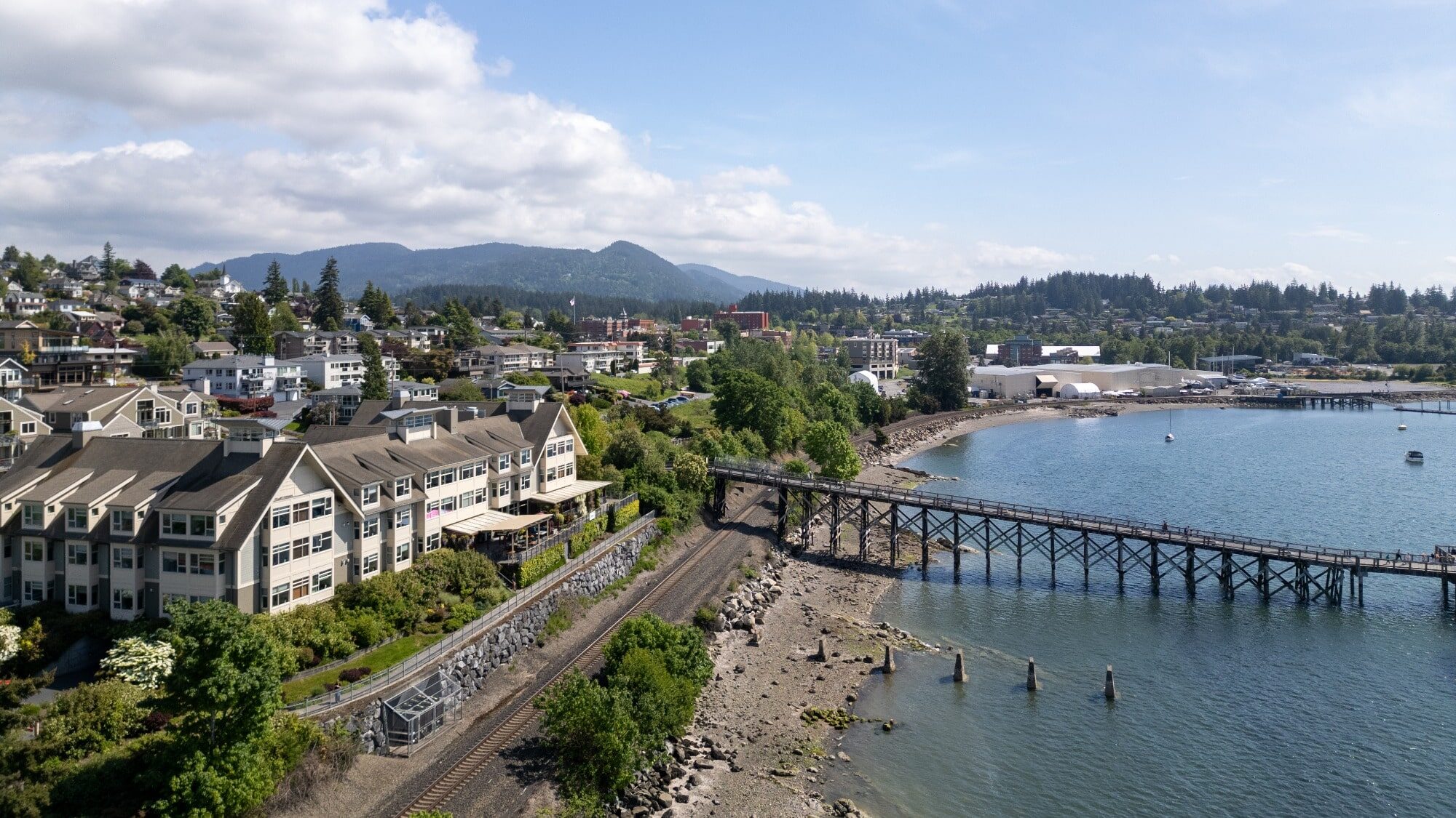 A coastal town in Washington with houses, a wooden pier, railway tracks, shoreline, and forested hills in the background under a partly cloudy sky offers a sense of renew and tranquility.