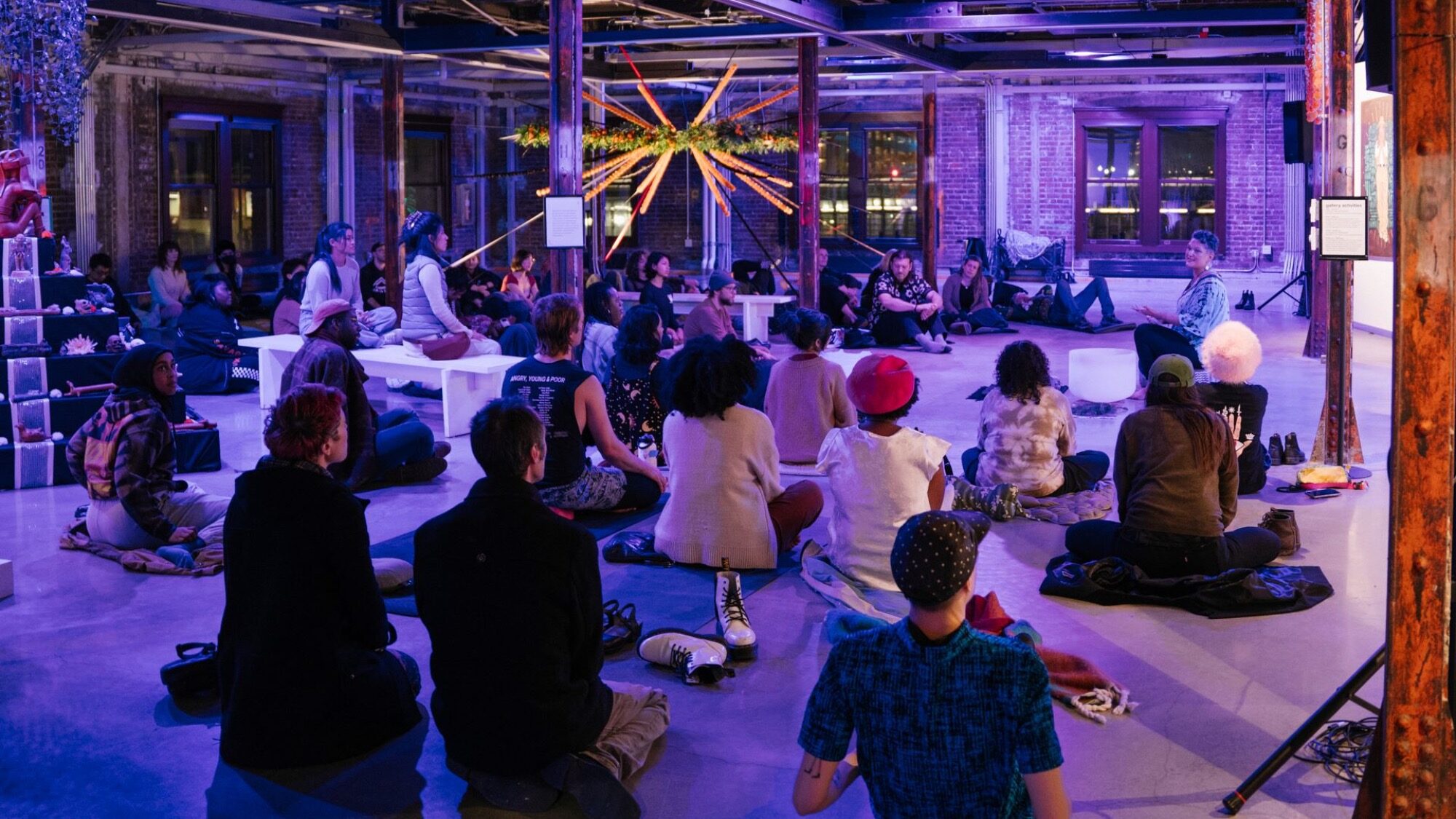A group of people sit on the floor and benches in a spacious, industrial-style room with exposed brick and colorful lighting, attentively facing a speaker.