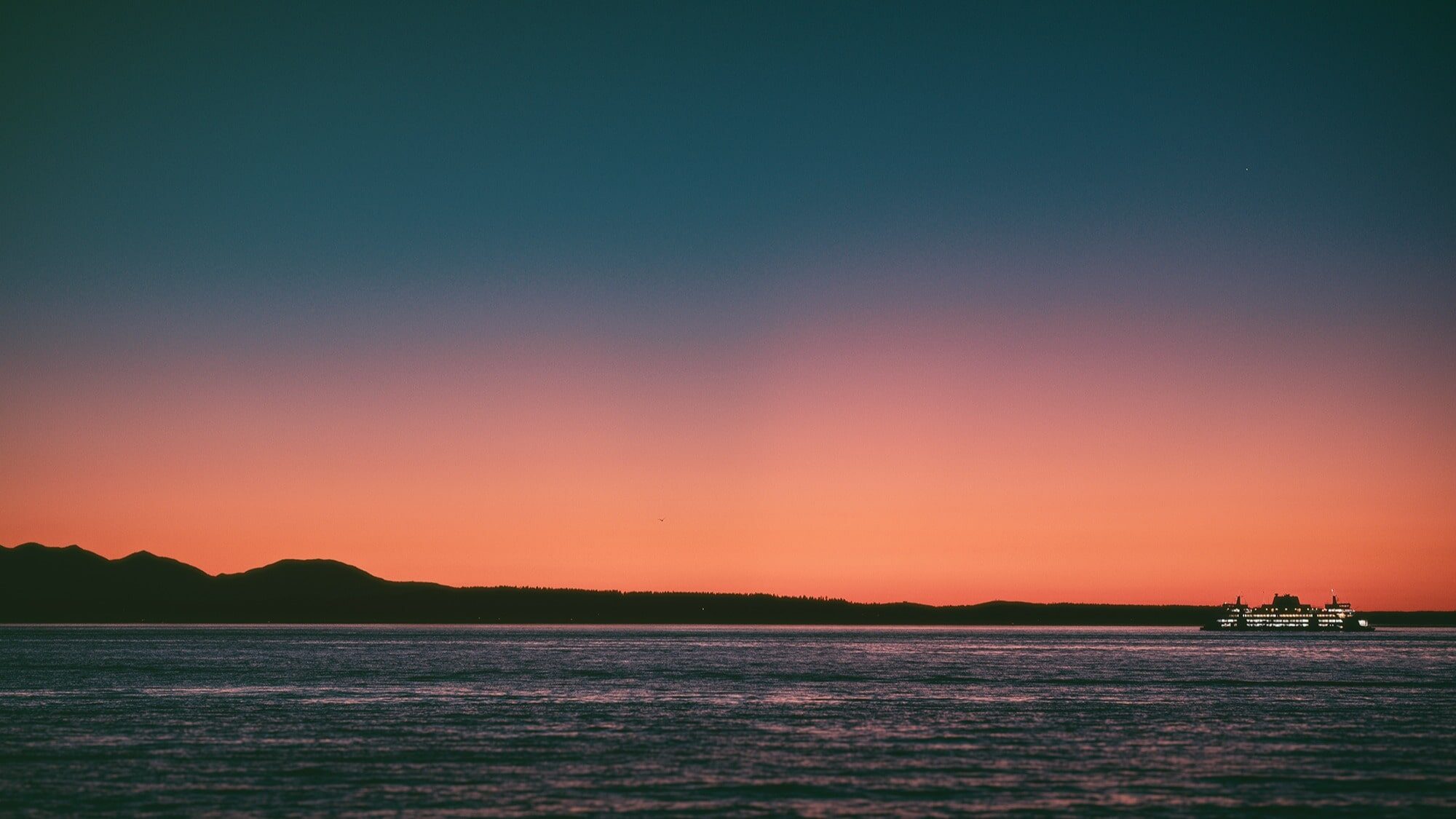 A ferry crosses calm water at sunset with mountains silhouetted in the background and a gradient sky from orange to deep blue, capturing a moment of tranquility perfect for photography enthusiasts like Nick Ward.
