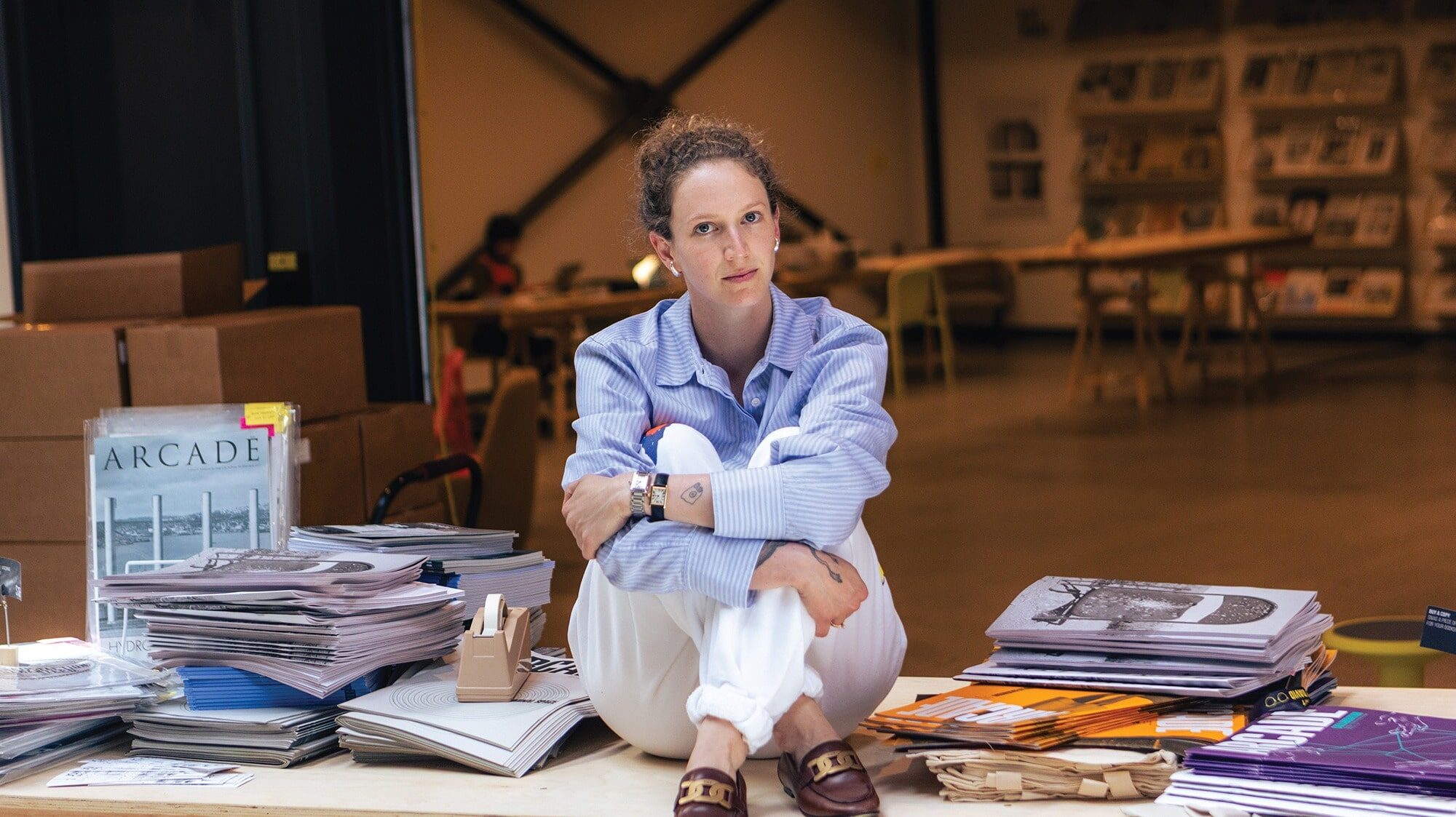 A person sits cross-legged on a table surrounded by stacks of magazines and papers in a modern indoor space, immersing themselves in architecture and design with the latest issue of ARCADE.