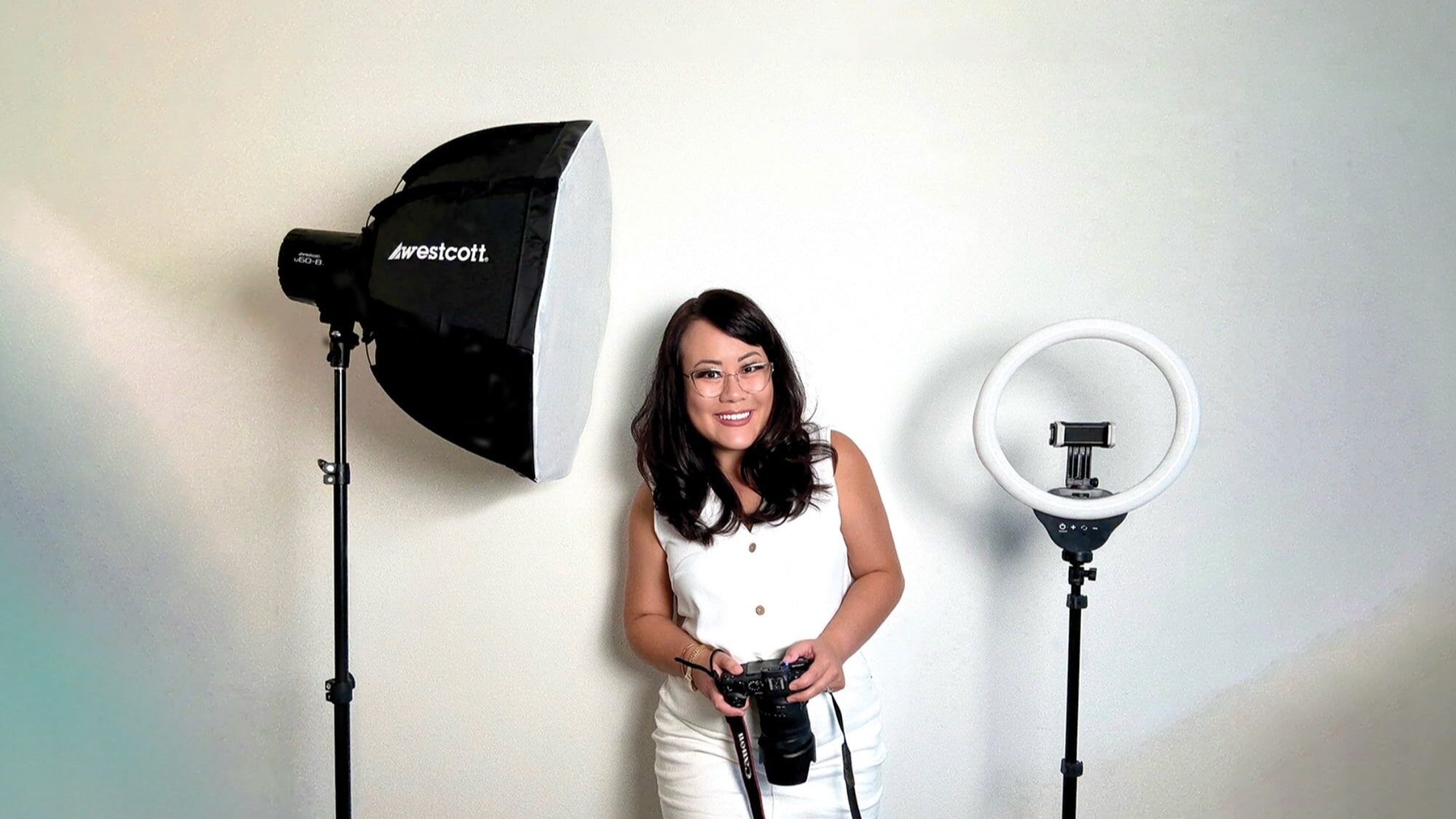 A woman in white stands between a studio softbox light and a ring light, holding a camera with a sense of future thinking, in front of a plain white wall.