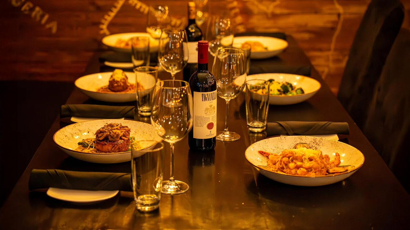 A dining table set for four with plates of pasta dishes, a bottle of Sansonina red wine, wine glasses, and black napkins, in a warmly lit Ristorante Italiano setting.