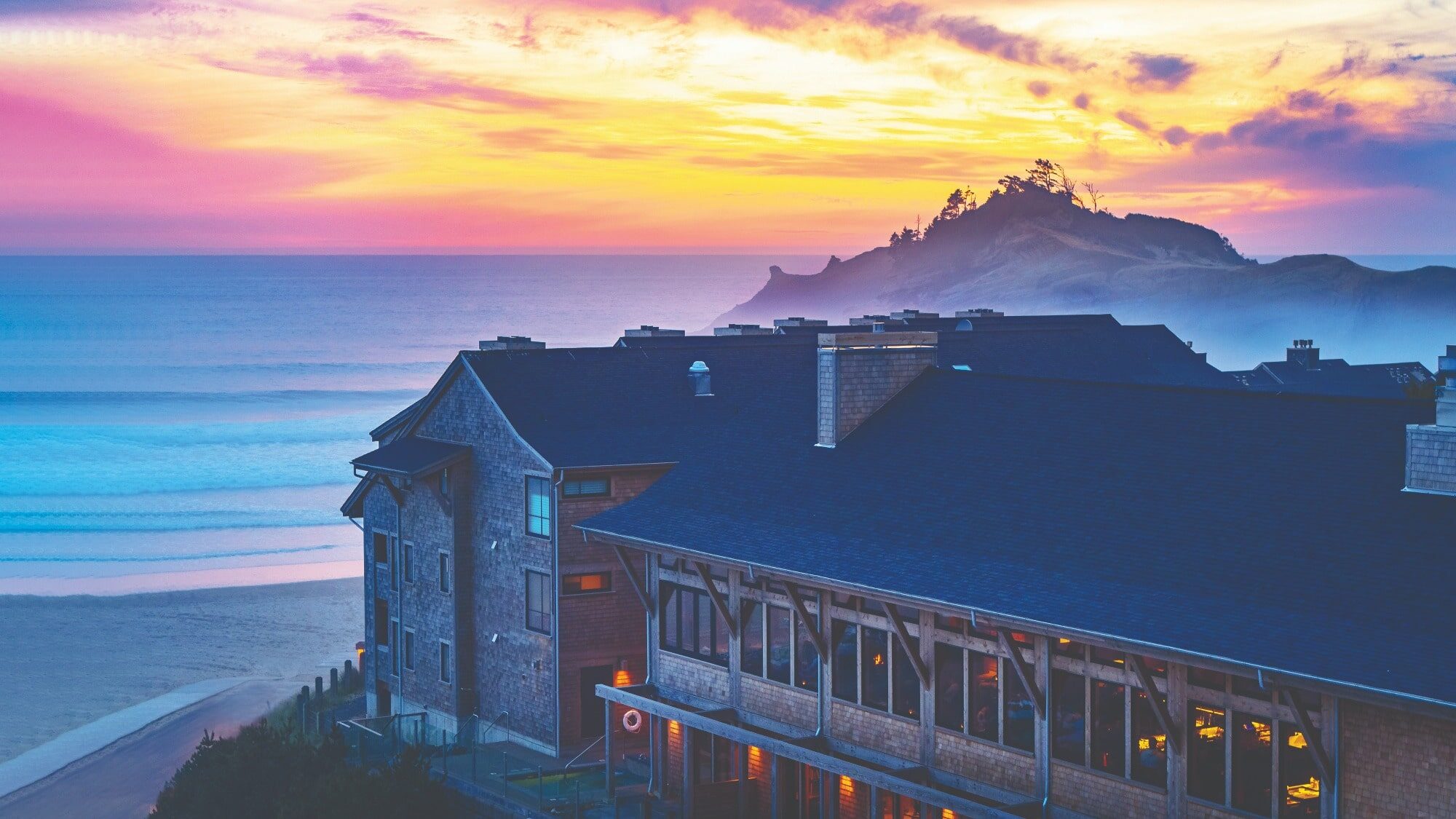A coastal hotel overlooks the ocean at sunset, offering beautiful scenery with waves, a sandy beach, and a rocky, tree-covered headland in the background under a colorful West Coast sky.