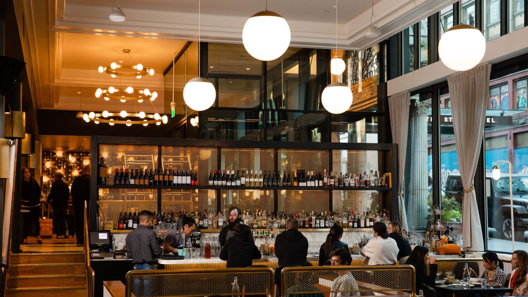 Restaurant bar area with patrons seated at the counter and tables, bartenders working, wine bottles displayed on shelves, and large globe lights overhead.