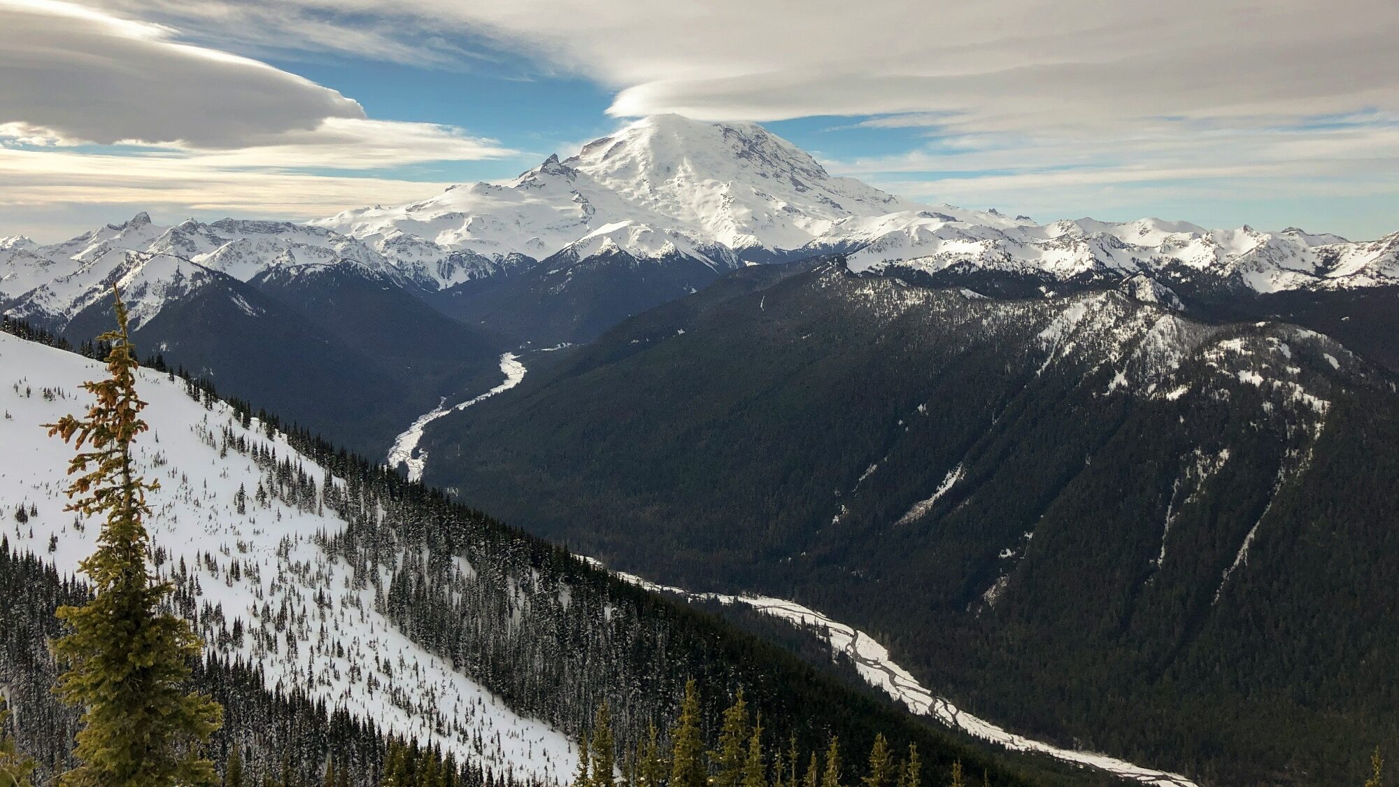 Snow-capped mountain with a dramatic cloud above, surrounded by Northwest forested hills and a winding river in a deep valley—nature’s beauty, even as winter approaches.