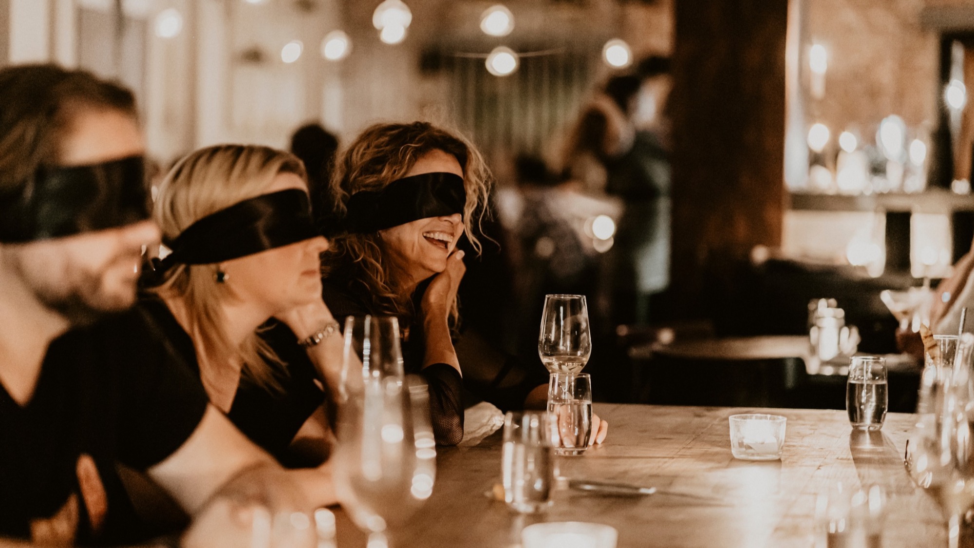 Three people sit at a candlelit table wearing blindfolds, with empty wine glasses and water glasses in front of them, in a dimly lit restaurant setting.