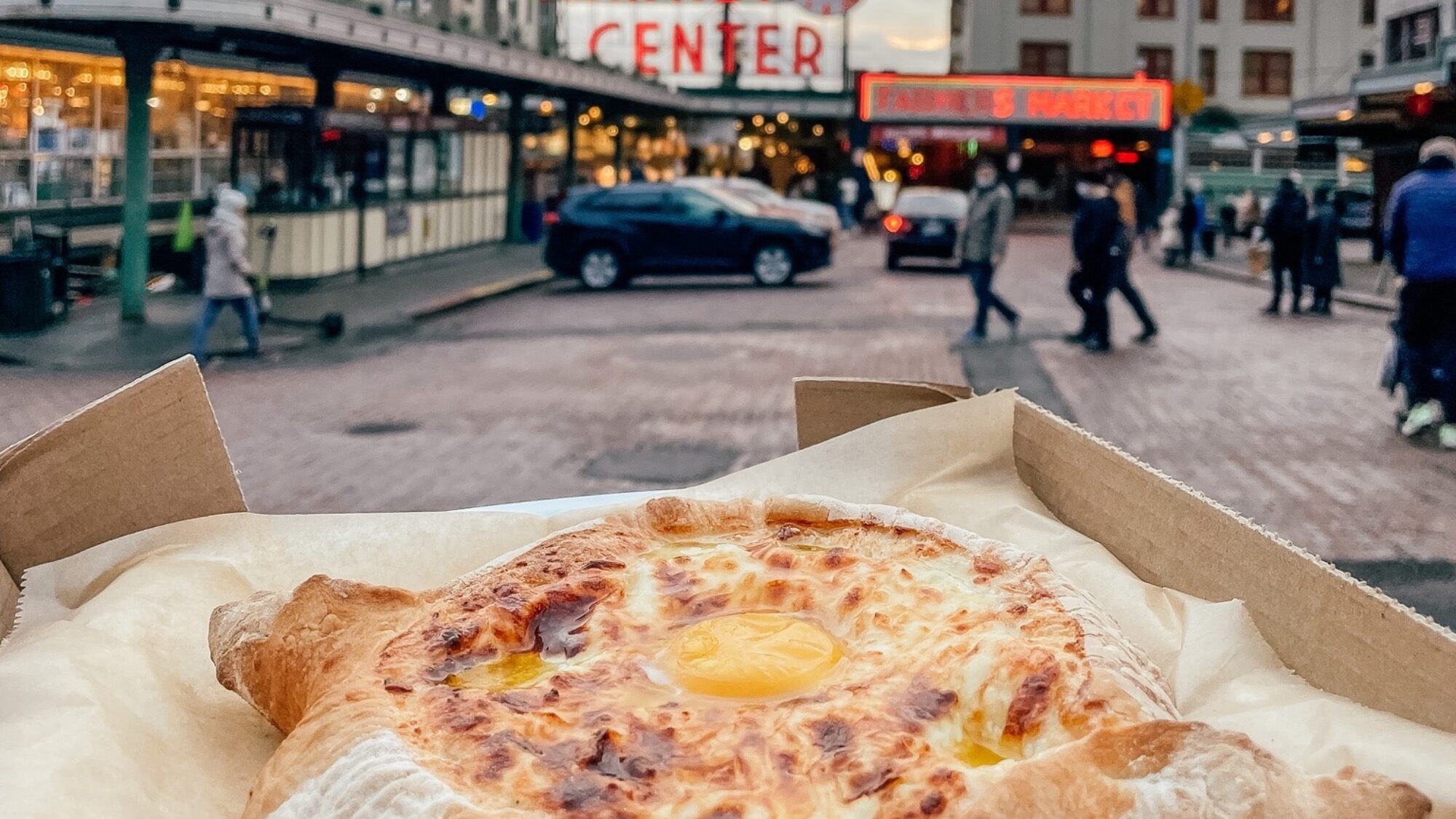 A close-up of a baked bread dish with melted cheese and egg in the center, held in a tray outdoors near a busy market street.