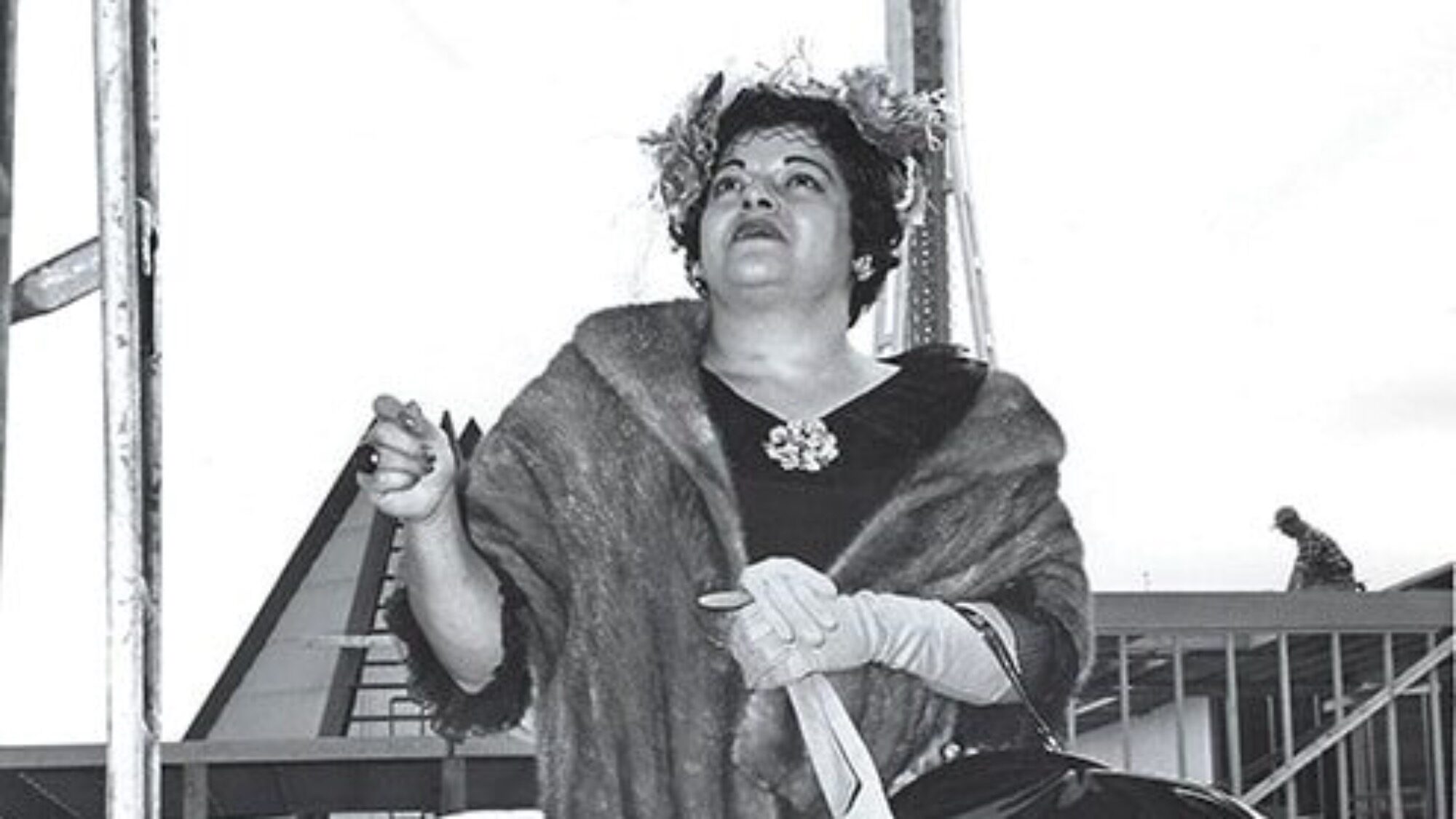 A woman in a fur shawl and gloves stands outdoors, holding a handbag and umbrella, with a building and staircase in the background.