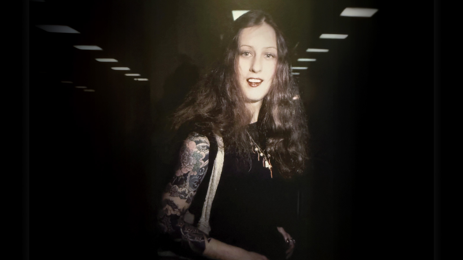 A person with long brown hair, a tattooed arm—an Ink Queen—wearing a black top stands indoors under ceiling lights, looking at the camera and smiling slightly.