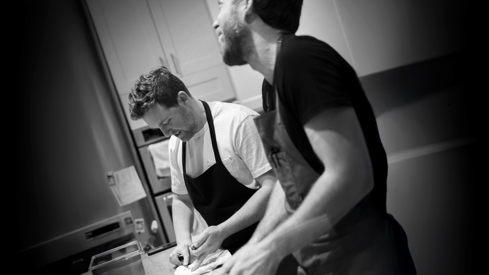 Two men in aprons prepare food together in a kitchen, one slicing ingredients while the other smiles and talks. The image is in black and white.