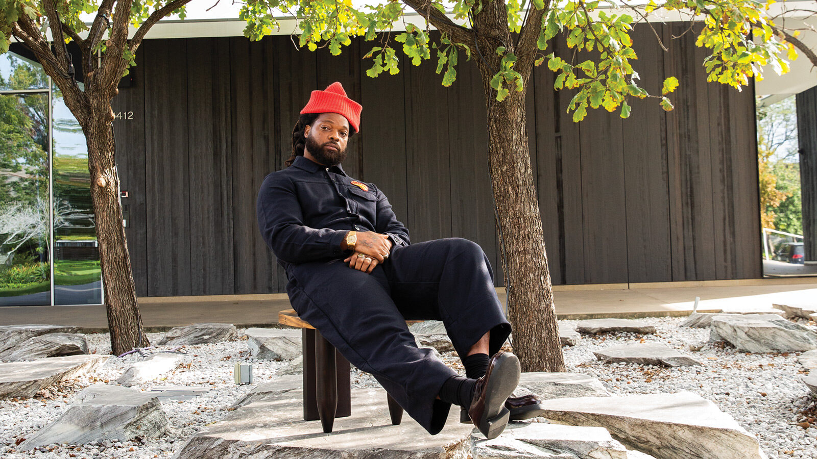 A person in a navy jumpsuit and red hat sits on a bench outdoors, surrounded by trees and rocks, in front of a modern building—giving off Design Star vibes reminiscent of Michael Bennett’s signature style.