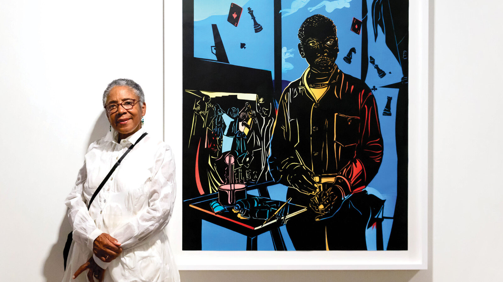 An older woman in a white outfit, Cultural Torchbearer Barbara Earl Thomas, stands next to a colorful framed painting of a person at a table, displayed on a gallery wall.