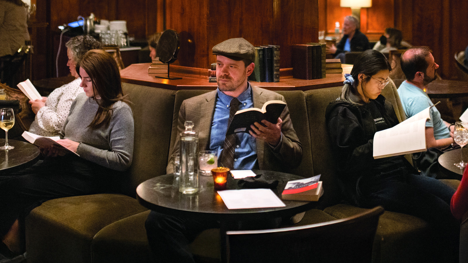 Several people sit on couches in a dimly lit lounge, each reading books—an atmosphere reminiscent of a Literary Leader event hosted by Christopher Frizzelle—with drinks and a candle on the tables.