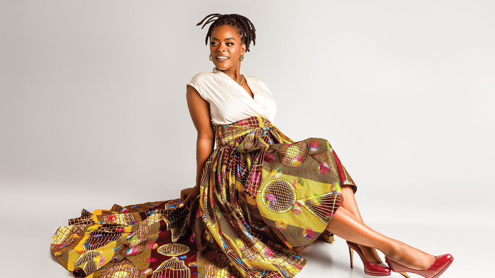 Kiesha B. Free, also known as The Connector, sits gracefully on the floor in a cream top and colorful patterned skirt with a long train, paired with striking red high heels against a plain white background.