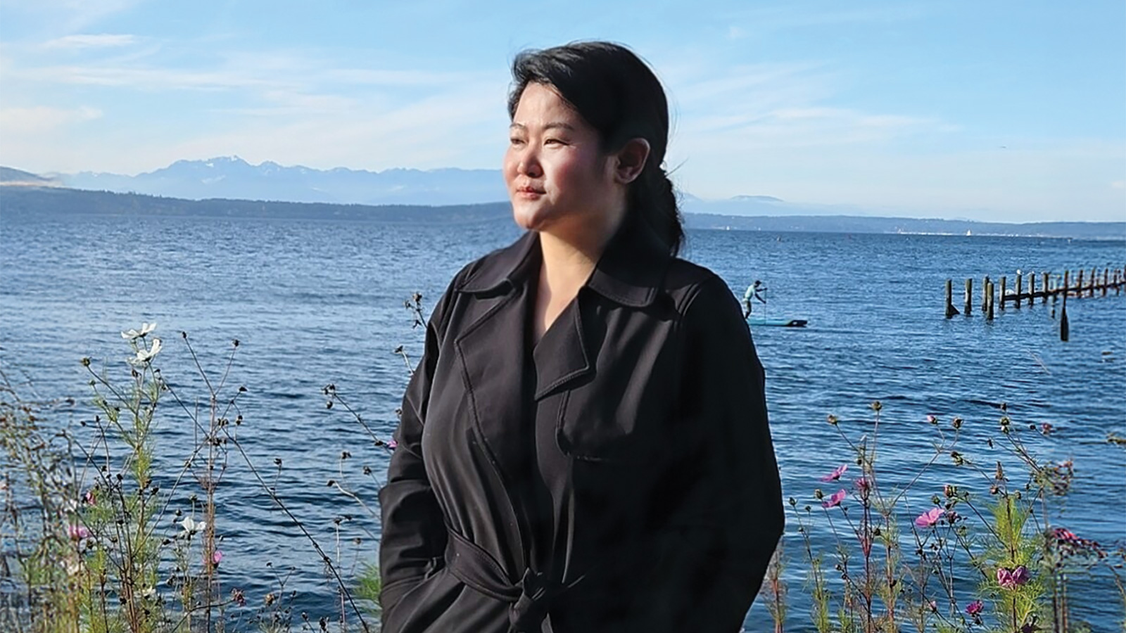 A person in a dark coat stands by a lake with wildflowers in the foreground, mountains in the distance, and a blue sky above—restaurateur Lily Wu finds inspiration for her next restaurant amid this tranquil scene.