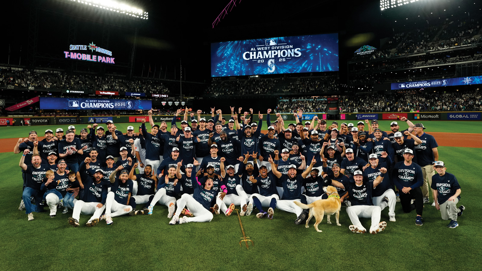 The Mariners baseball team celebrates on the field at night in front of an "AL West Division Champions" sign at T-Mobile Park, with fans in the stands behind them—true Boys of Summer making history.