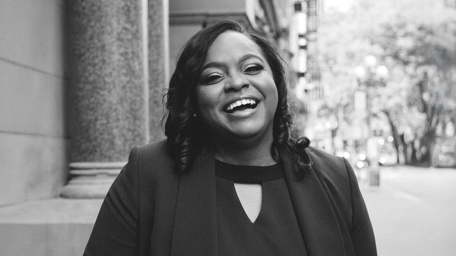 Black and white photo of Michelle Merriweather standing outdoors, smiling in a blazer with buildings and trees in the background, radiating positive influence.