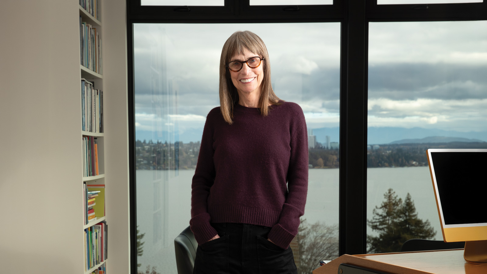 A woman with straight brown hair and glasses stands smiling in front of a large window overlooking water and trees—bookshelves and a computer nearby—embodying the spirit of a Big Giver, much like Shari D. Behnke.