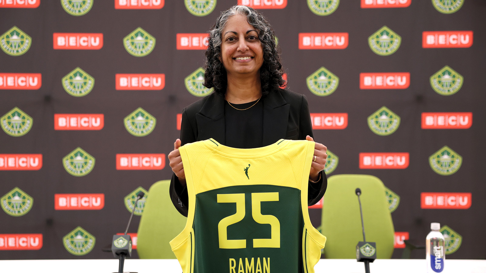 Sonia Raman, The Coach, stands holding a yellow and green basketball jersey with the name "RAMAN" and number 25, against a backdrop with Seattle Storm and sponsor logos.